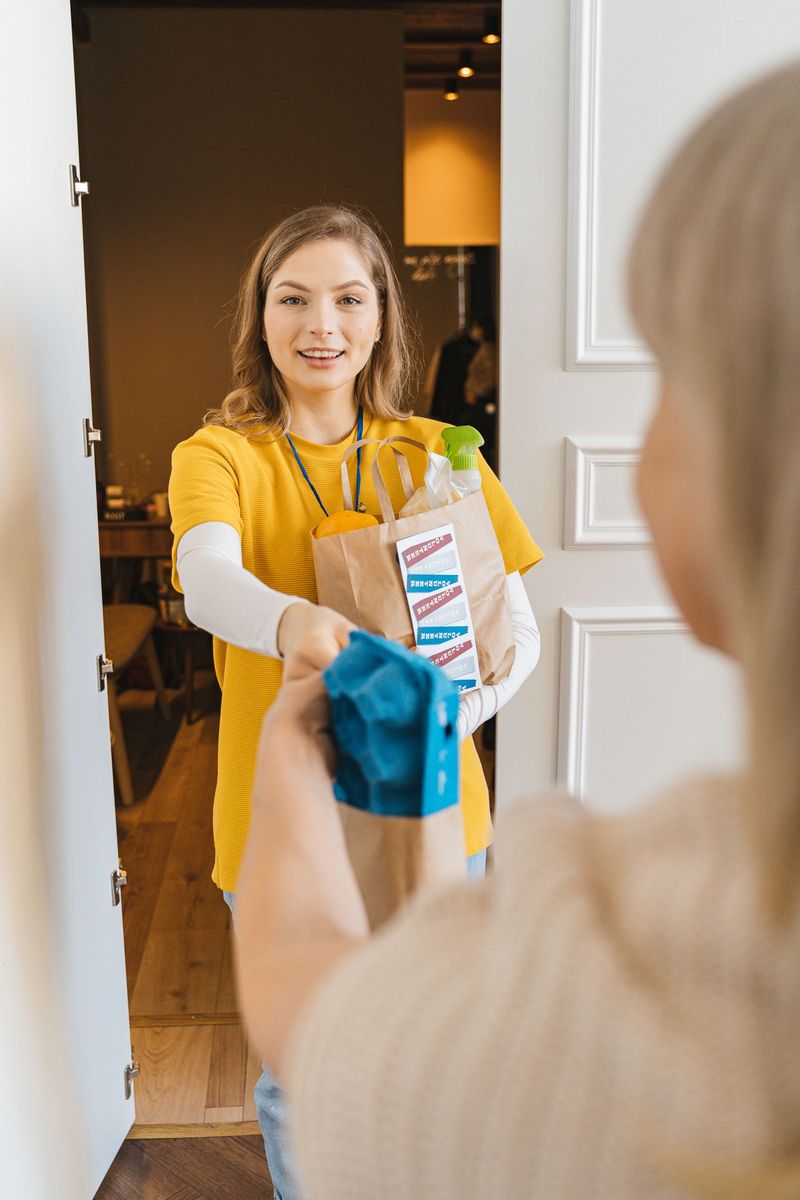 A smiling woman in a yellow shirt delivering a paper bag of groceries to a neighbor at the door